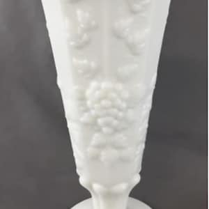 May include: A tall, white, vintage-style vase with a flared top and a round base. The vase features a textured, embossed pattern of grapes and leaves. The vase is made of opaque white glass and stands approximately 10 inches tall.