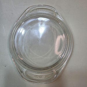 May include: Clear glass Pyrex oval casserole dish lid with two handles. The lid is transparent, allowing a view of the contents. The design is simple and functional, suitable for oven use. The lid is a replacement part.