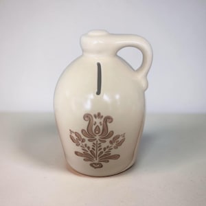 May include: Cream-colored ceramic jug with a handle and a decorative floral design in brown. A vertical black line is painted on the front. The jug has a rounded body and a small spout.