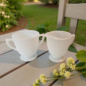 May include: Two white, vintage-style milk glass cream and sugar set. The creamer has a handle, and the sugar bowl has two handles. Both pieces have a ribbed design and sit on a pedestal base. The set is displayed on a gray wooden surface.