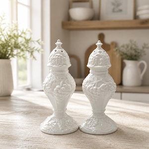 May include: A pair of white ceramic salt and pepper shakers featuring a raised relief pattern of grapes and leaves. Each shaker has a tapered body, a wide circular base, and a removable domed top with small holes for dispensing seasonings.