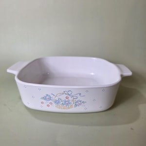 May include: White square baking dish with a floral basket design in blue, pink, and green. The dish has two handles and is suitable for oven use. A classic piece for baking and serving.