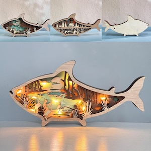 May include: A wooden shark-shaped night light with a detailed scene of a lake with trees and birds inside. The light is illuminated with warm white LED lights.