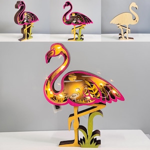 Puede incluir: Escultura de flamenco de madera con un diseño en capas, con colores rosa, amarillo y marrón. La escultura tiene un diseño recortado detallado con una cadena de luces en el interior. El flamenco está sobre una base verde y amarilla.