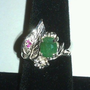 May include: A silver ring with a green gemstone and pink accents. The ring features a leaf design with intricate details.