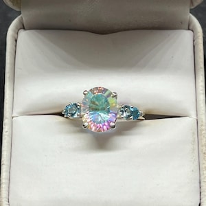 May include: A silver ring featuring a large, oval-cut, iridescent mystic topaz center stone with a rainbow-like sheen, flanked by two smaller, round-cut, light blue gemstones on each side, all set in a polished silver metal band.