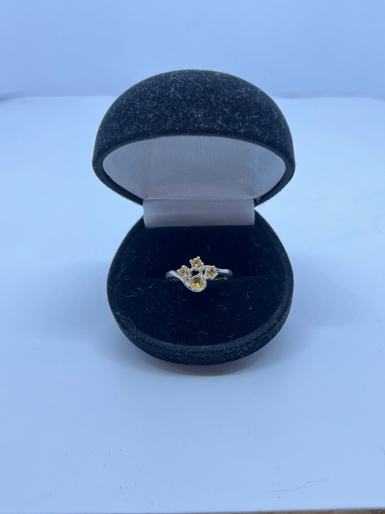 May include: A silver ring with a cluster of five yellow gemstones set in a curved design. The ring is displayed in a black velvet ring box.