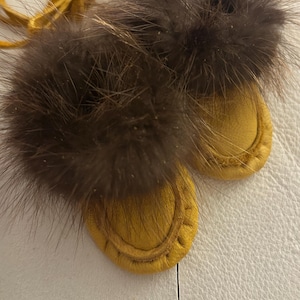 Könnte beinhalten: Ein Paar Babymokassins mit einer Basis aus senfgelbem Leder und dunkelbraunem Pelzbesatz. Die Mokassins sind mit einer senfgelben Lederschnur zusammengebunden. Nahaufnahme.