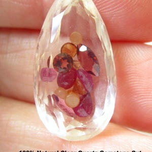 May include: A pear-shaped clear quartz gemstone pendant with a hollowed center filled with loose ruby, pink and orange sapphire, red and orange garnet, and peach moonstone. The text reads: 100% Natural Clear Quartz Gemstone Cut, Hallowed out Then Filled With Ruby, Pink & Orange Sapphire, Red & Orange Garnet, Peach Moonstone. Drilled As Shown.