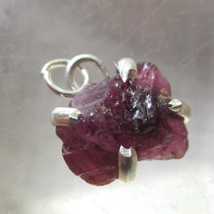 May include: A raw, natural ruby pendant with a silver bail. The ruby is a deep, rich pink color and has a rough, textured surface. The silver bail is simple and elegant, with a loop for a chain.