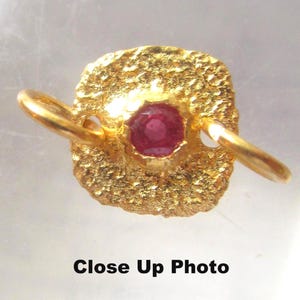 May include: Close-up of a gold-colored pendant with a textured, square-shaped design. A round, red gemstone is set in the center. The pendant has two gold loops on either side. The text "Close Up Photo" is at the bottom.