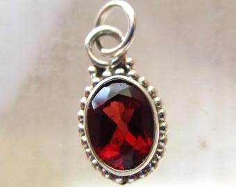 May include: A silver pendant featuring a faceted, oval-shaped, deep red gemstone. The gemstone is set in a beaded bezel and has a silver loop for a chain. The pendant is a piece of jewelry.