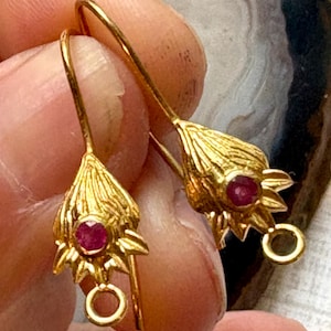 May include: A pair of gold-colored earrings featuring a floral design with a small, round, red gemstone at the center of each flower. The earrings have a hook closure and are displayed against a blurred background.