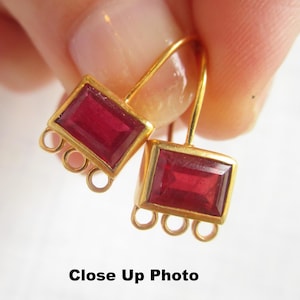 May include: Close-up of gold-colored earrings with rectangular, deep red gemstones. Each earring has a gold frame around the gemstone and three small gold loops below. The text "Close Up Photo" is at the bottom.