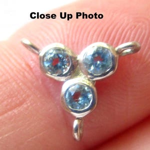 May include: A close-up photo of a silver pendant with three light blue gemstones. The gemstones have a circular shape with a red center. The pendant has a loop at the bottom and two loops on the sides. The text "Close Up Photo" is at the top.