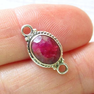 May include: An oval-shaped, faceted deep pink ruby gemstone set in a silver-toned metal frame with a twisted rope border. The setting features two small circular loops on opposite ends, allowing it to function as a jewelry connector or link.