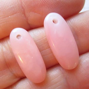 May include: Two pale pink, oval-shaped beads with a hole at the top for stringing. The beads have a smooth, glossy finish. These beads are ideal for jewelry making.