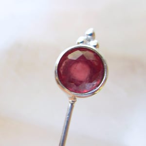 May include: A silver stick pin featuring a round, faceted deep red gemstone set in a bezel. The top of the setting is accented with three small, smooth silver spheres arranged in a triangular cluster above the circular stone mount.