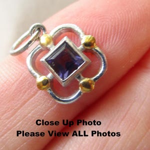 May include: A square-shaped pendant featuring a central deep purple gemstone set in silver. The frame has a four-lobed design with small gold-toned spheres at each corner and a silver jump ring attached at the top for hanging on a chain.