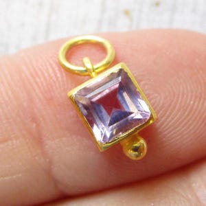 May include: A gold-colored square pendant with a light purple gemstone. The pendant has a small gold loop at the top and a small gold ball at the bottom. The gemstone is square-cut and reflects light.