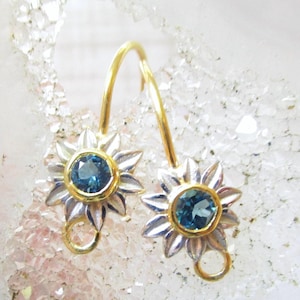 Könnte beinhalten: Ein Paar gold- und silberfarbene Ohrringe mit einem runden blauen Edelstein in einer Goldfassung. Das Design ähnelt einer Blume mit silbernen Blütenblättern und einer kleinen goldenen Öse unten, befestigt an einem geschwungenen Goldhaken.