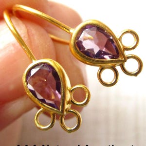 May include: A pair of gold-colored earrings with teardrop-shaped, purple gemstones. Each earring has a loop at the top and three small loops at the bottom. The earrings are held in a person's hand.
