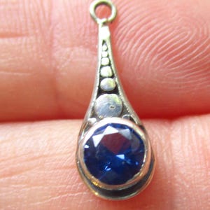 May include: A silver-toned metal pendant featuring a teardrop-shaped frame with a series of graduated circular accents leading to a single, round, faceted deep blue gemstone set at the base, complete with a small circular jump ring at the top.