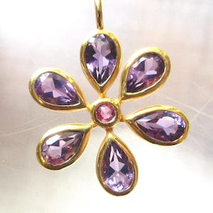 May include: A flower-shaped pendant featuring five pear-cut purple amethyst petals surrounding a central round pink ruby, set in 18K gold over 925 sterling silver. Text reads: Natural Ruby & Amethyst Gemstones 18K Gold Over 925 Sterling Silver.