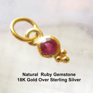 Könnte beinhalten: Ein goldfarbener Anhänger mit einem runden, facettierten roten Rubin in einer kreisförmigen Fassung, akzentuiert durch drei kleine Goldkugeln an der Basis und einen Biegering oben. Der Text lautet: Natural Ruby Gemstone 18K Gold Over Sterling Silver.