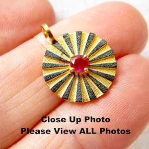Może przedstawiać: Złoty wisiorek z czarno-złotym wzorem promieni słońca i czerwonym kamieniem szlachetnym w środku.