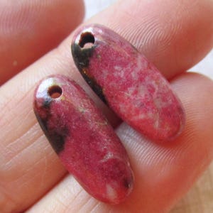 Peut inclure: Deux pendentifs en pierre précieuse de thulite naturelle. Les pendentifs sont d'un rose vif avec des accents brun foncé et un trou pour l'enfilage. Le texte indique "100% Natural Thulite Gemstone Sold Per Pair."
