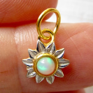 May include: A small flower-shaped pendant featuring a central round white opal cabochon set in a gold bezel. The petals are crafted from silver-toned metal with a textured finish. A gold jump ring is attached to the top of the floral design.