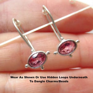 May include: A pair of silver-toned earrings featuring oval-shaped, faceted pink stones set in metal frames with small decorative prongs. The text "Wear As Shown Or Use Hidden Loops Underneath To Dangle Charms/Beads" is printed at the bottom of the image.