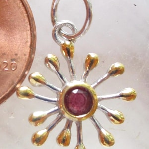 May include: A silver and gold-toned pendant featuring a central round red gemstone surrounded by twelve radiating spokes with gold-toned tips. A silver jump ring is attached to the top. A copper-colored US penny is visible for scale.