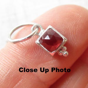 May include: A close-up of a small, silver-colored charm with a square, faceted, deep red gemstone. The charm has a silver loop for attachment and a cluster of three small silver beads. The text "Close Up Photo" is visible.