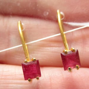 May include: A pair of gold-colored earrings with rectangular, deep red gemstones. The earrings have a simple, elegant design, with a small gold hook and four small gold accents around each gemstone. The gemstones are a vibrant, eye-catching color.