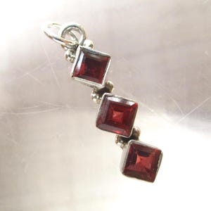 May include: A silver pendant featuring three square-cut, deep red garnet gemstones arranged in a vertical line. The stones are set in silver bezels with small silver bead accents between each square setting and a silver jump ring at the top.