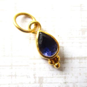 May include: A gold-toned pendant featuring a teardrop-shaped dark blue gemstone set in a smooth bezel. The pendant includes a circular jump ring at the top for attachment and a small decorative cluster of three tiny gold spheres at the base.