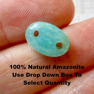 Op de afbeelding: Een lichtblauwe, ovale amazoniet kraal met twee gaten. De kraal wordt in een hand gehouden. De tekst op de afbeelding luidt "100% Natural Amazonite".