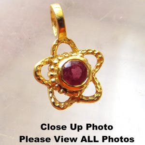 Może przedstawiać: Złoty wisiorek w kształcie kwiatu z czerwonym kamieniem szlachetnym w środku.