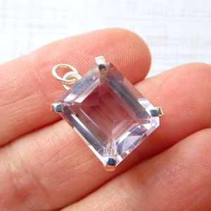May include: A faceted, rectangular pendant with a light purple hue. The gemstone is set in a silver-colored metal frame with a small loop at the top for a chain. The pendant is held in a hand.