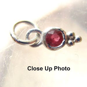 May include: A close-up of a silver-toned pendant with a round, faceted, red gemstone. The pendant has a loop for a chain, a knot design, and three small silver beads. The text "Close Up Photo" is at the bottom.
