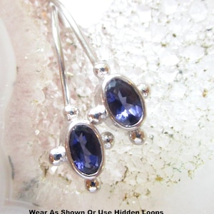 May include: A pair of silver earrings featuring oval-cut dark blue stones set in metal frames with small spherical accents. The text at the bottom reads: Wear As Shown Or Use Hidden Loops Underneath To Dangle Beads/Charms. The design includes hooks.
