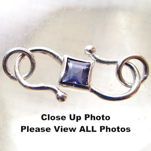 May include: Close-up of a silver clasp with a square, dark blue gemstone at its center. The clasp has a decorative, curved design with a small ball at the end of the hook. The text "Close Up Photo Please View ALL Photos" is at the bottom.