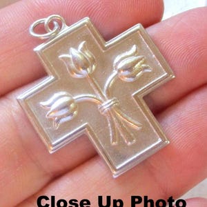 May include: Silver-toned cross pendant with a bouquet of tulips in the center. The cross has a detailed design with a loop at the top for a chain. The text "Close Up Photo" is at the bottom.