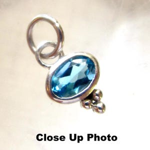 May include: A silver pendant featuring an oval-cut blue gemstone set in a smooth metal bezel. The pendant includes a circular jump ring at the top and a decorative cluster of three small silver spheres at the bottom. The text Close Up Photo is visible.