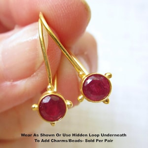 May include: A pair of gold-tone earrings featuring round, faceted deep red stones set in metal bezels with four small decorative prongs. The earrings include a curved wire hook and a small loop underneath. Text reads: Wear As Shown Or Use Hidden Loop Underneath To Add Charms/Beads- Sold Per Pair
