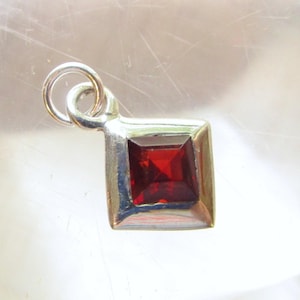 May include: A silver-toned pendant featuring a square-cut, deep red gemstone set in a smooth metal frame. The pendant includes a small circular jump ring at the top for attachment to a chain, showcasing a polished finish and a faceted stone surface.