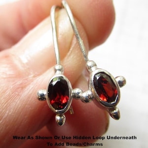 May include: A pair of silver drop earrings featuring oval-cut deep red garnet stones set in metal frames with small spherical accents. The text on the image reads: Wear As Shown Or Use Hidden Loop Underneath To Add Beads/Charms Sold As Pair.