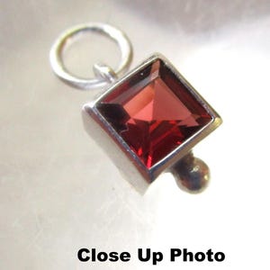 May include: A silver charm featuring a square red gemstone. The charm has a loop at the top for attaching to a bracelet or necklace.